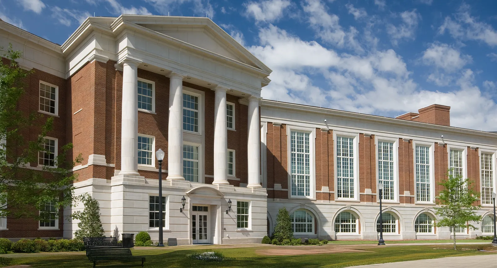 A landscape image of a brick and mortar university with a bench, trees and grass in front.