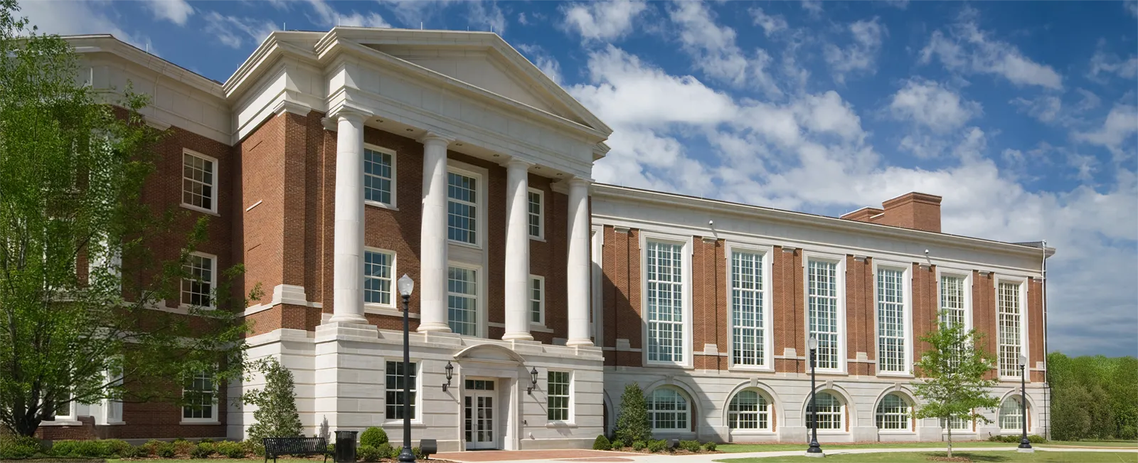 A wide photo of grass trees circumscribing a brick and mortar university with tall windows and high columns.